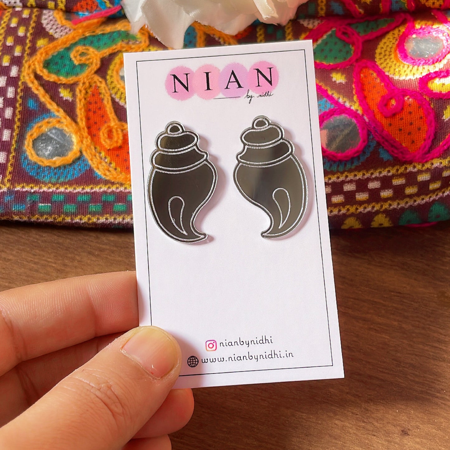 Shubh Shankh Earrings - Nian by Nidhi - Glassy Silver - placed in a brown colorful background, held in a hand