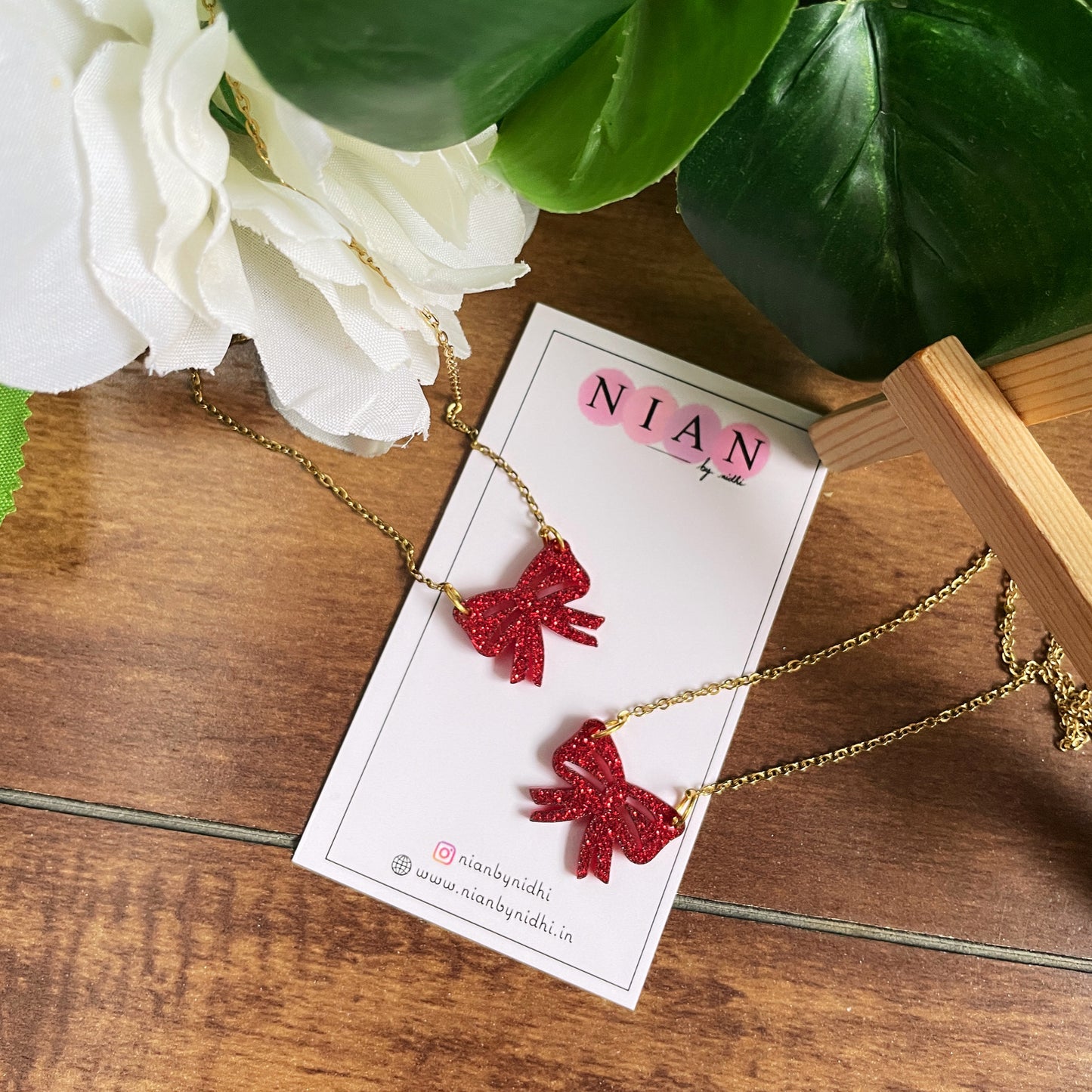 2 pieces of Bow Necklace - Shimmer Red - Nian by Nidhi - in a white, brown, and green background