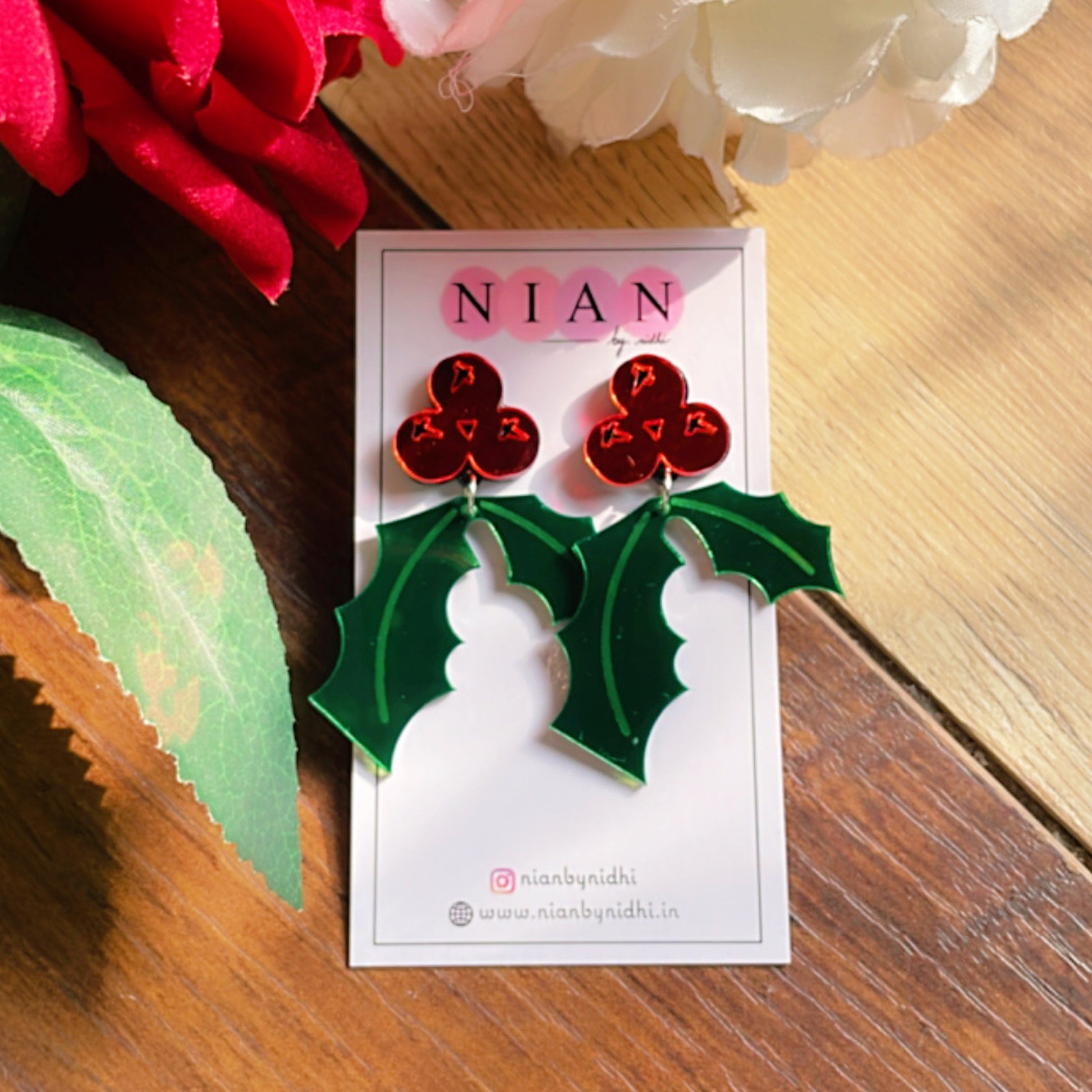 Holly Leaf Earrings - Green and Red- Nian by Nidhi- placed in a brown and flowery background