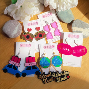 Bon Voyage Combo (Set of 5) - consists 5 multi-colored earrings - Happy Journey Earrings, Wanderlust Earrings, Camera Earrings, Be Happy Be Sassy Earrings, and Glam Goggles Earrings - Nian by Nidhi - placed in a brown background