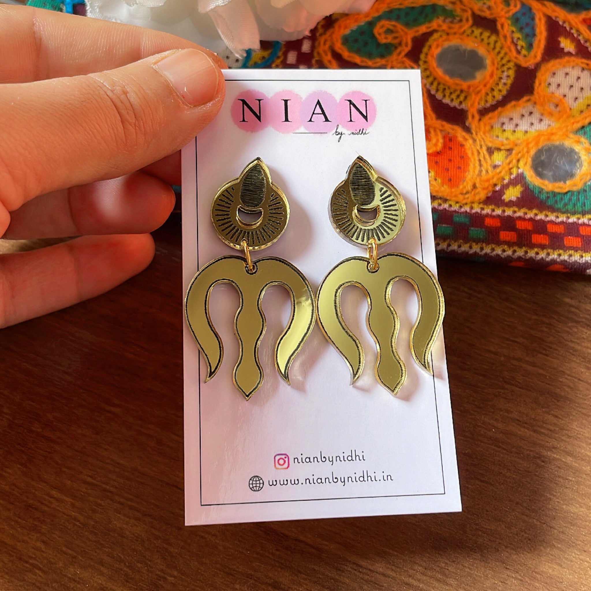 Trishul Earrings - Nian by Nidhi - Glassy Golden - placed in a brown and colorful background, held in a hand
