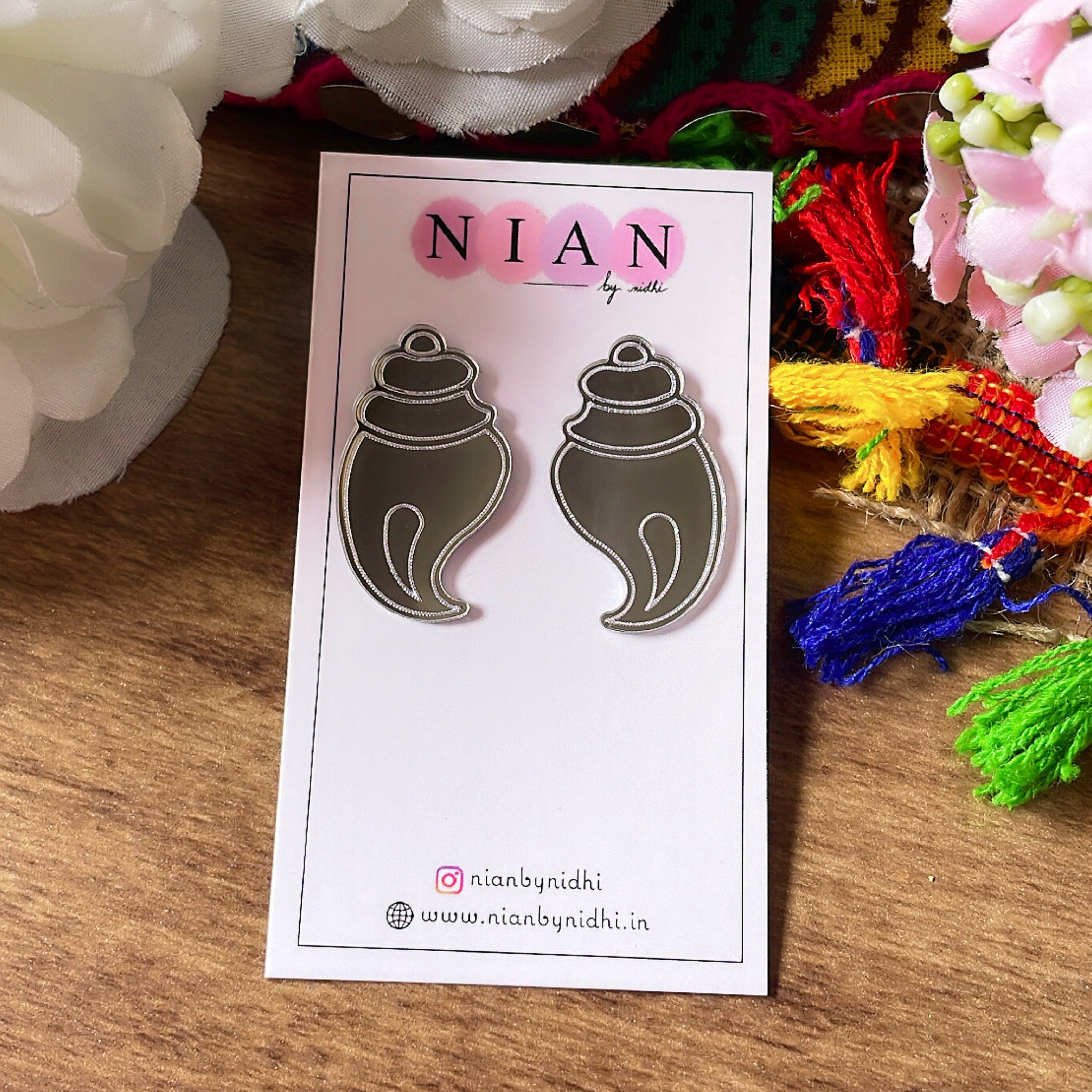 Shubh Shankh Earrings - Nian by Nidhi - Glassy Silver - placed in a brown colorful background