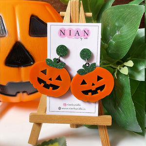 Plumpy Pumpkin Earrings - Orange, Black and Shimmer Green - Nian by Nidhi - placed on a small wooden canvas in a background that contains a halloween pumpkin and some leaves
