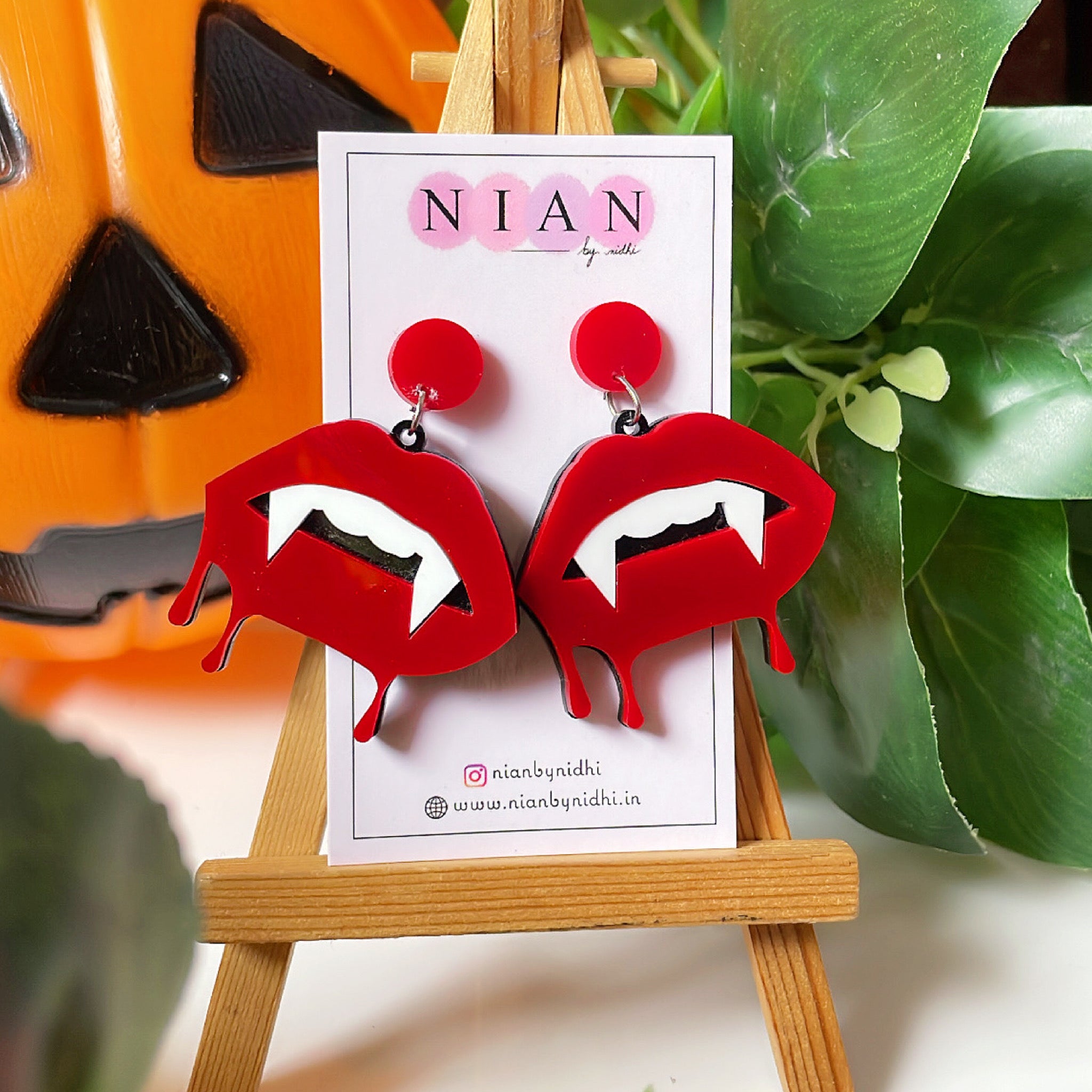 Bloody Lips Earrings - Red and White - Nian by Nidhi - placed on a small wooden canvas in a background that contains a halloween pumpkin and some leaves