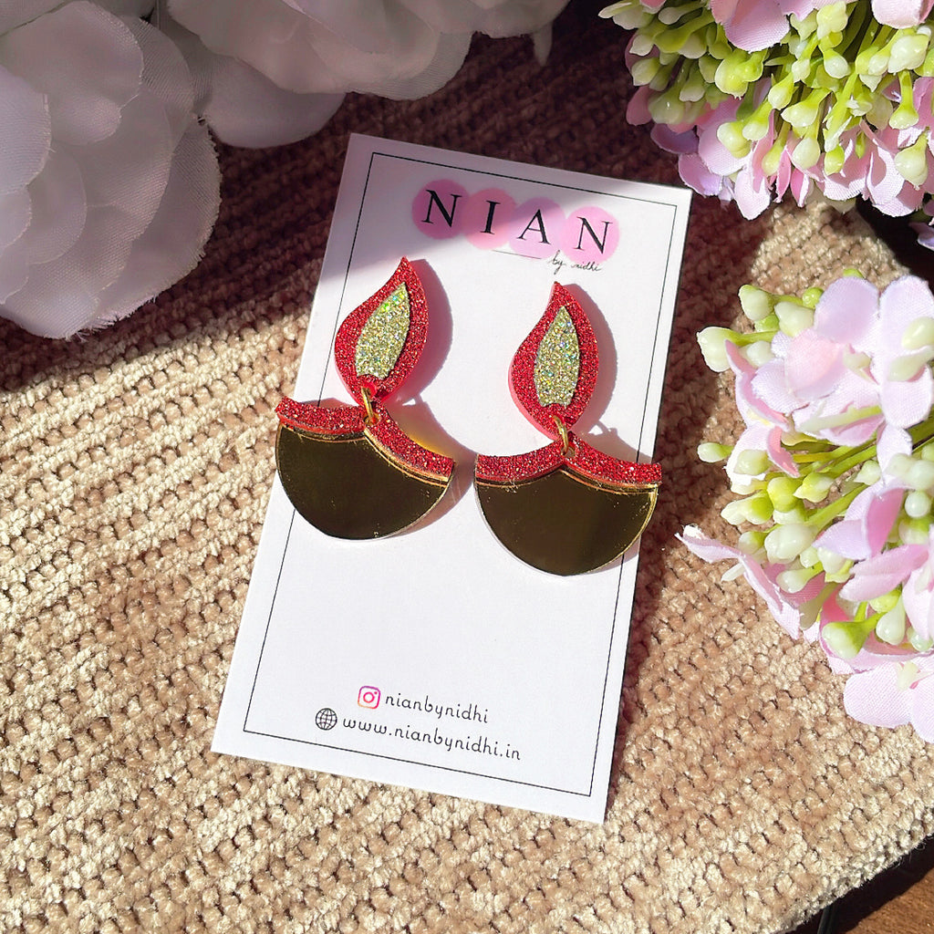 Dazzling Diya Earrings - Glossy Gold and Shimmer Red - Nian by Nidhi - placed in a light brown and a pink flowery background