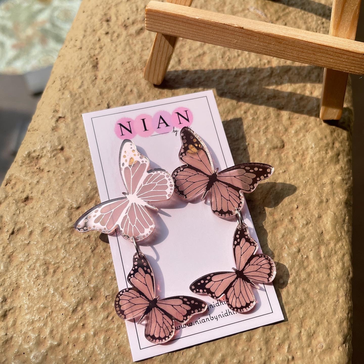 Butterfly Earrings - Glossy Rosegold - Nian by Nidhi - placed on a light brown surface, with a small wooden canvas in background