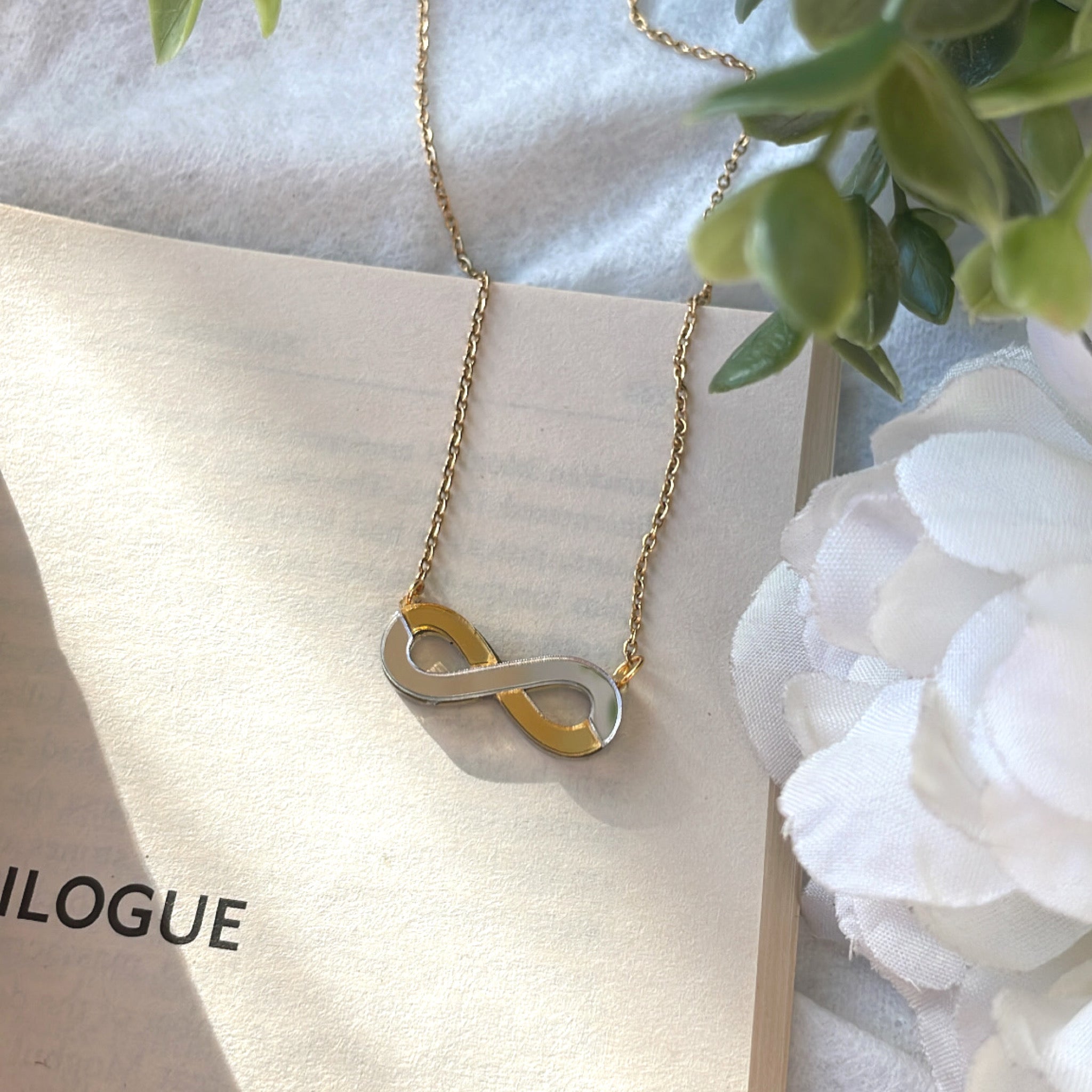 Infinity Necklace - Glossy Golden and Silver - Nian by Nidhi - placed in a white and green background, placed on a book