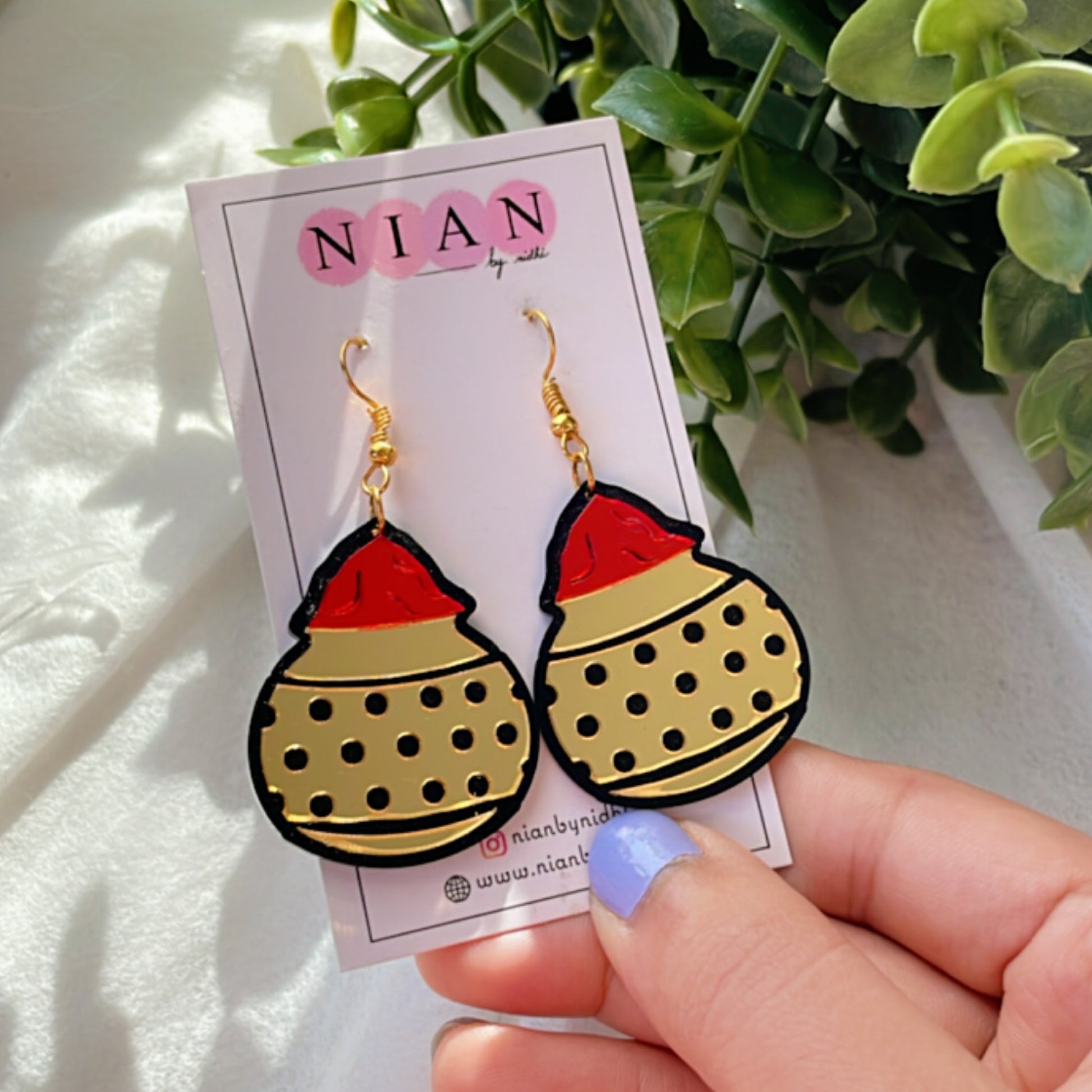 Gulal ki Matki Earrings - Glossy Golden and Red - Nian by Nidhi - placed in a white and green background, held in a woman's hand