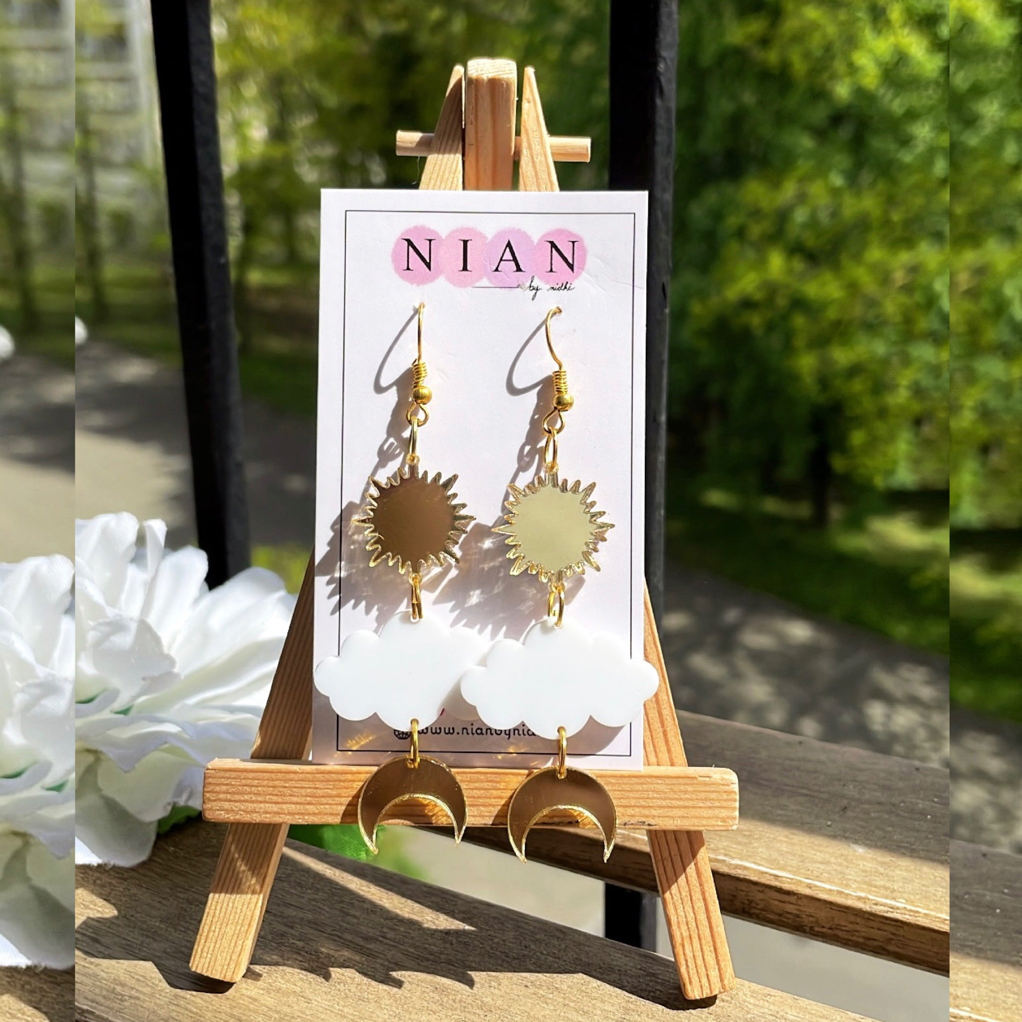 Shining Sky Danglers - Glossy Gold and White - Nian by Nidhi - placed on a small wooden canvas, with trees in the background