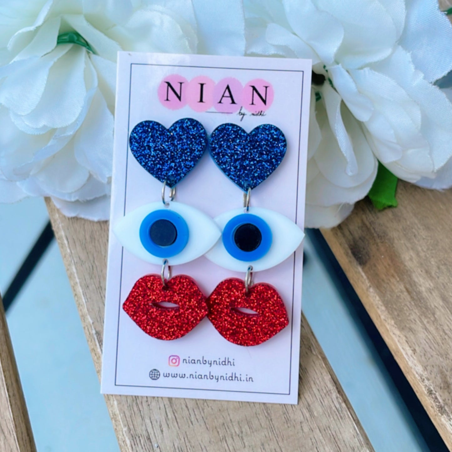 Evil Eye Charm Earrings - Shimmer blue, red and white - Nian by Nidhi - placed in a light beige background with a white flower