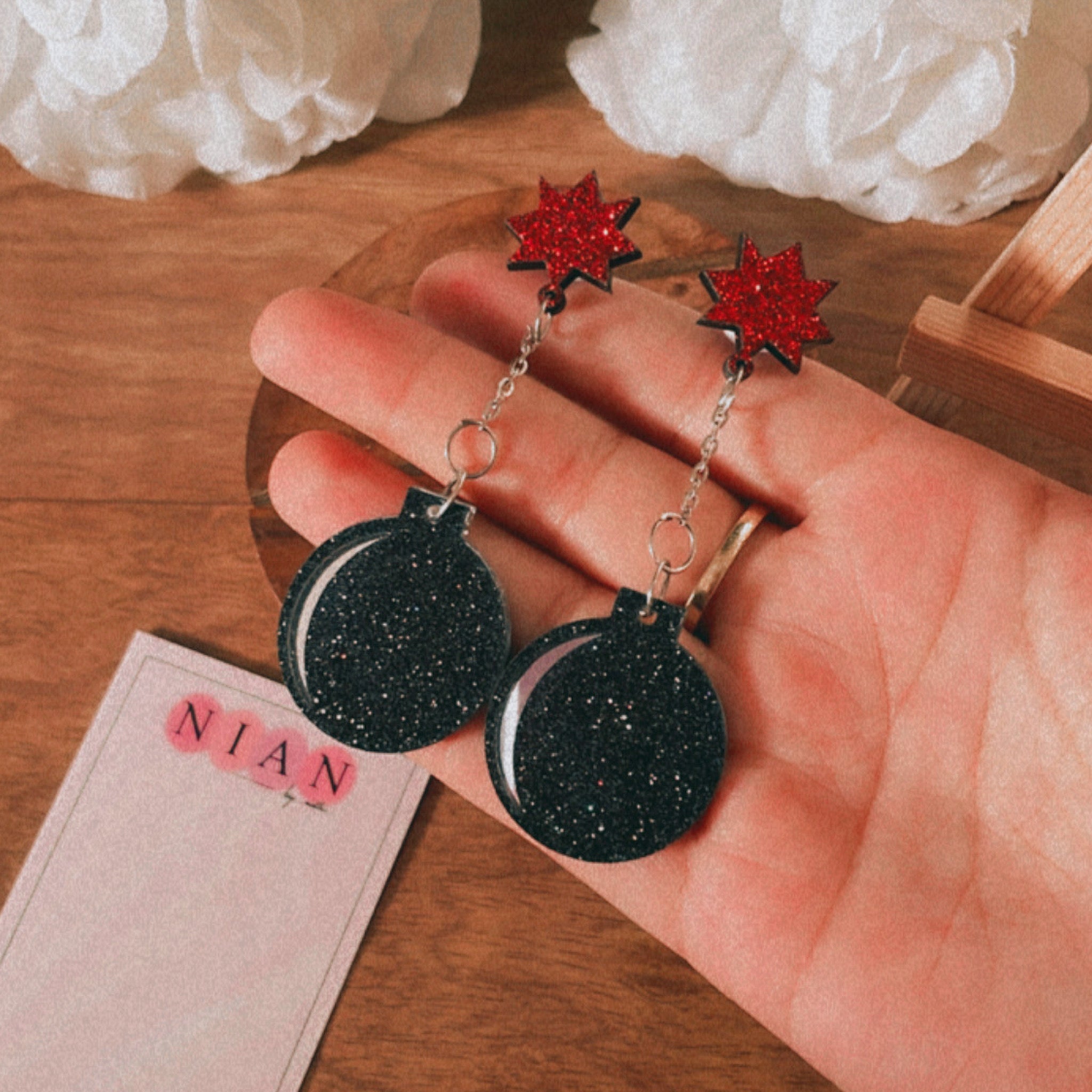 Pataka Earrings - Shimmer black and red - Nian by Nidhi - in a white and brown background, placed on a hand, with a Nian by Nidhi Earring card in the background