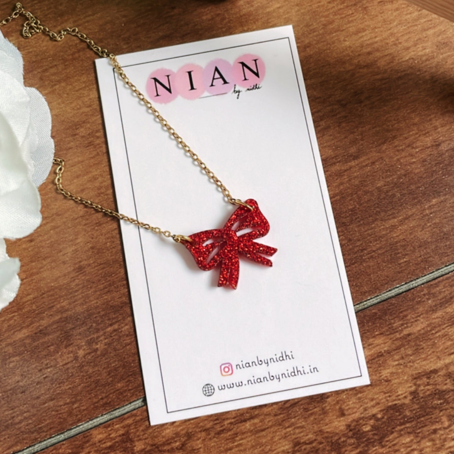 Bow Necklace - Shimmer Red - Nian by Nidhi - placed on a Nian by Nidhi Earring card, in a brown background