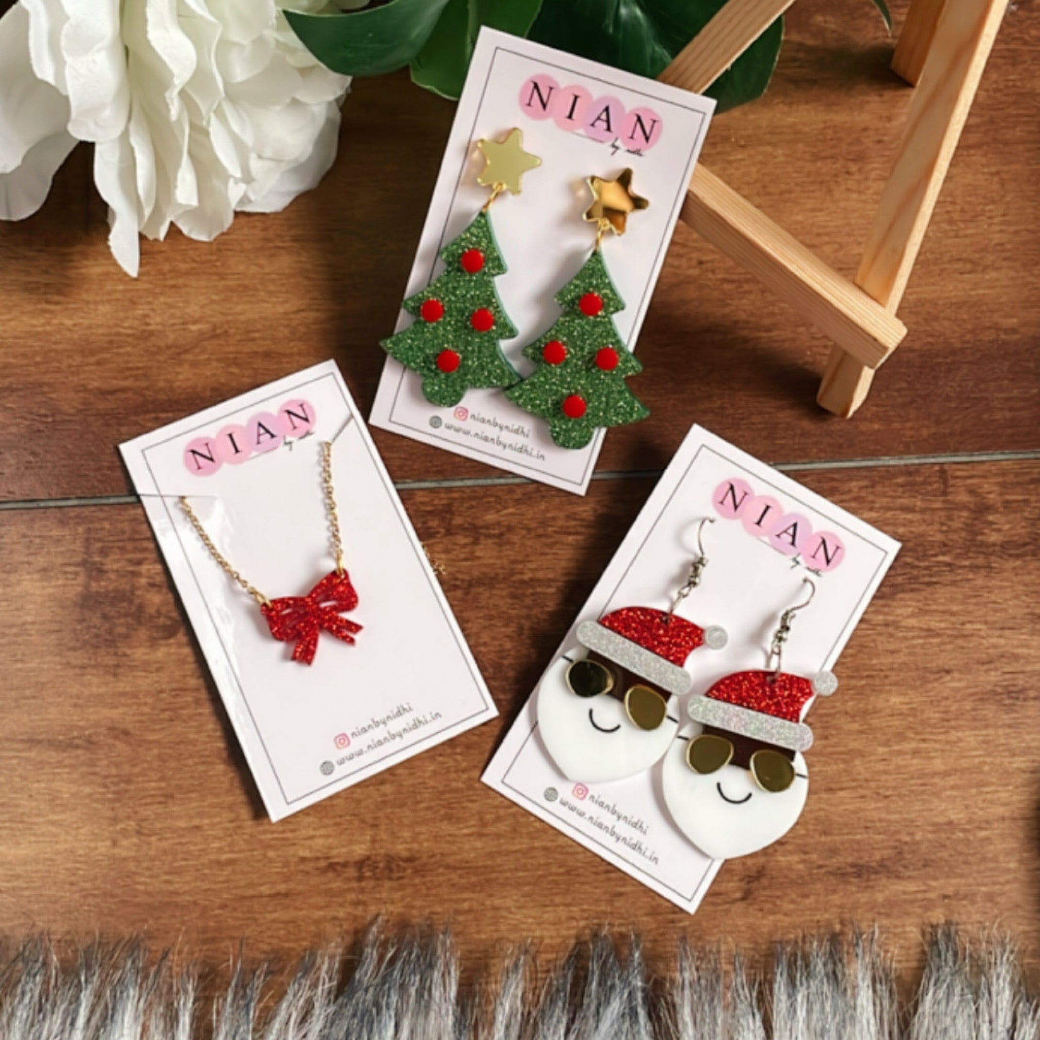 The Jingle Edition - consists 3 products - Yo Santa Earrings, Xmas Tree Earrings, and Bow Necklace - Nian by Nidhi - placed in a brown background along with Nian by Nidhi earring cards - Shimmer Green, Red, White, Shimmer Red