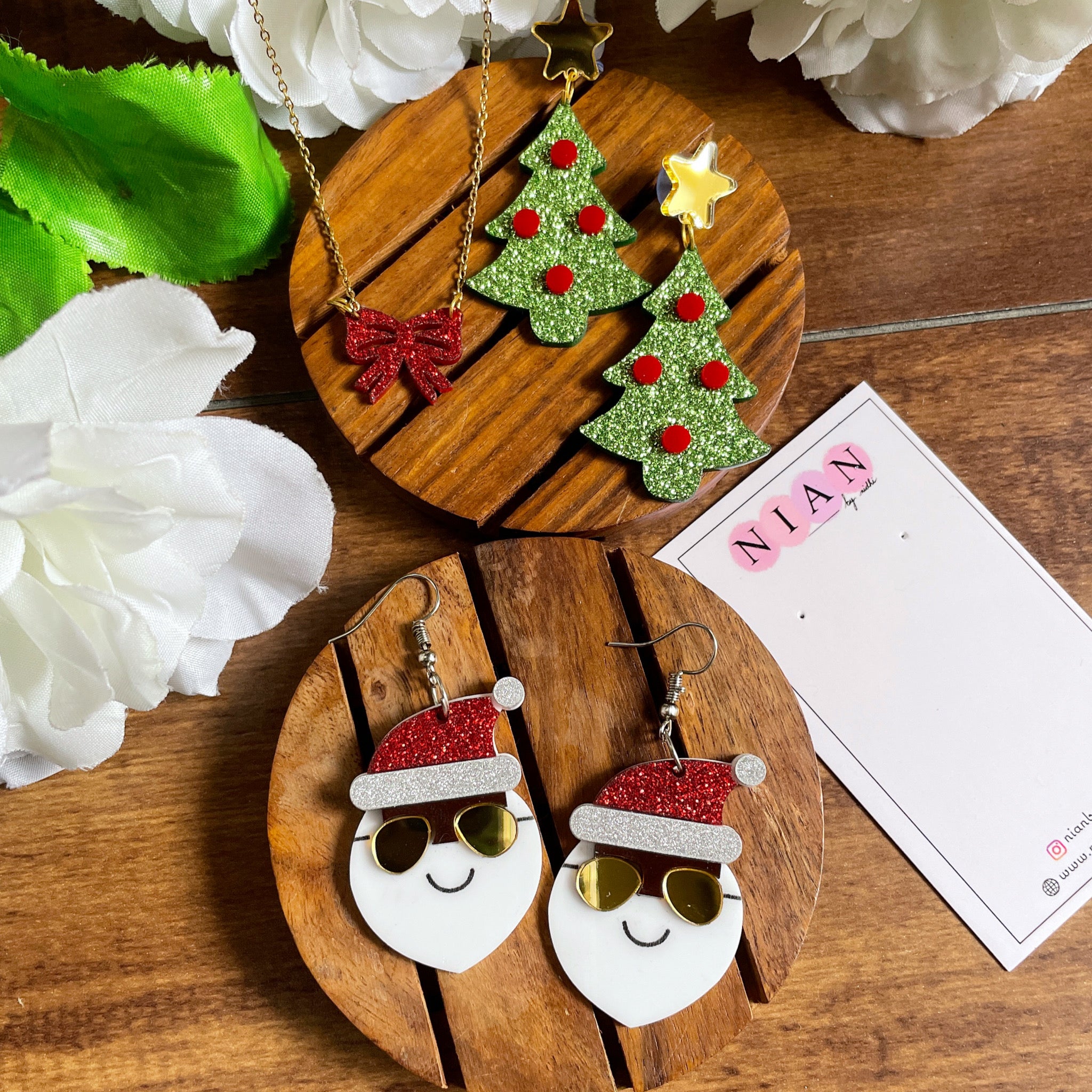 The Jingle Edition - consists 3 products - Yo Santa Earrings, Xmas Tree Earrings, and Bow Necklace - Nian by Nidhi - placed in a brown background along with a Nian by Nidhi earring card - Shimmer Green, Red, White, Shimmer Red