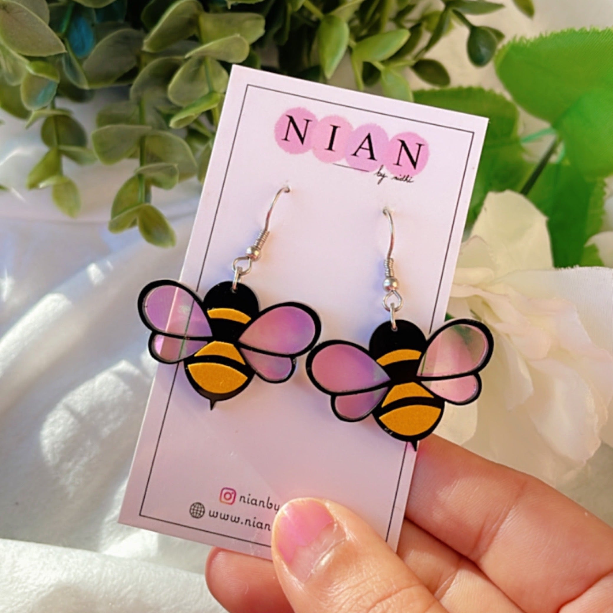 Baby Bee Earrings - Yellow and Black with a Holographin Effect - Nian by Nidhi - placed in a white and green background, with a Nian by Nidhi earring card
